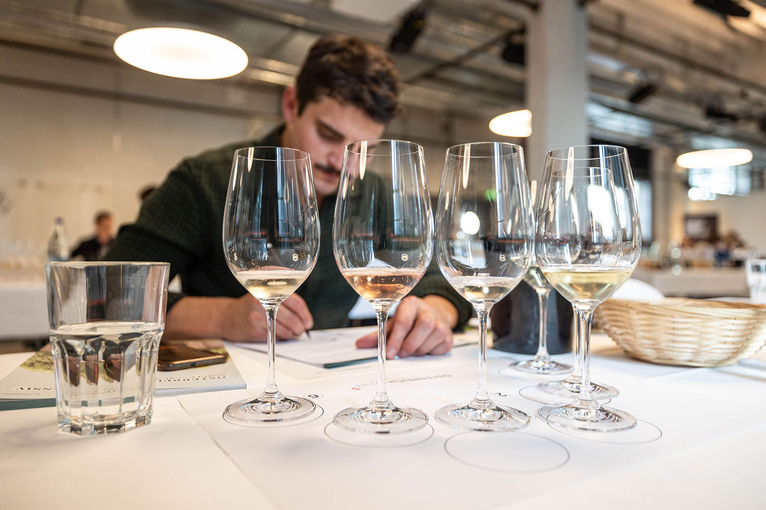 nahaufnahme von einem degustations set-up mit 6 weinen. im hintergrund ein junger herr, der seine notizen niederschreibt.